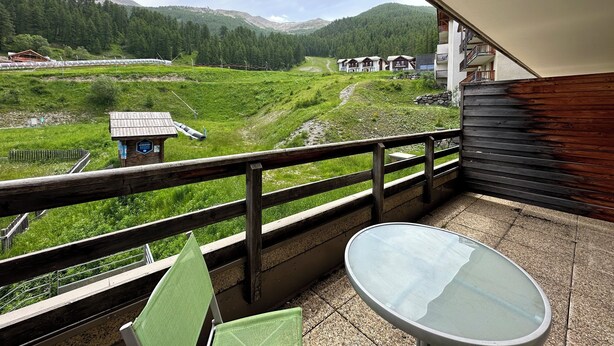 Een balkon met een glazen tafel en stoelen, met uitzicht op een bergachtig landschap met een klein gebouw en huizen in de verte.