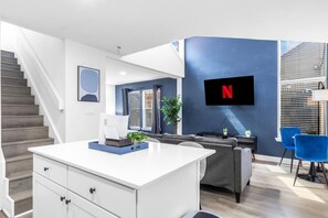 A modern living room with a white coffee table, grey sofa, and blue chairs. A flat-screen TV is mounted on the wall, and a staircase is visible in the background. - 5 Min to Love Field | Gated+Guard | 2BR | Parking (Dallas)