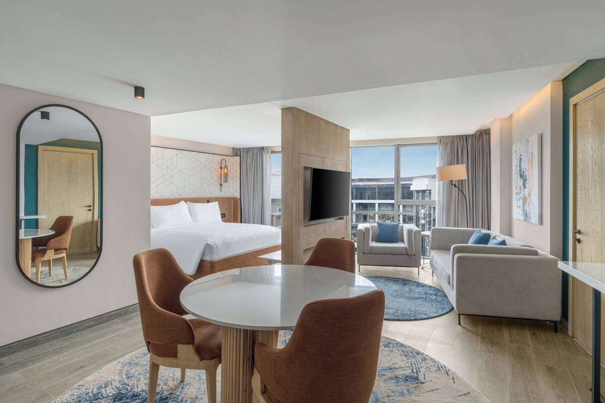 Une chambre d’hôtel comprenant un lit, une table à manger avec des chaises, un canapé, une télévision et une grande fenêtre donnant sur la ville.