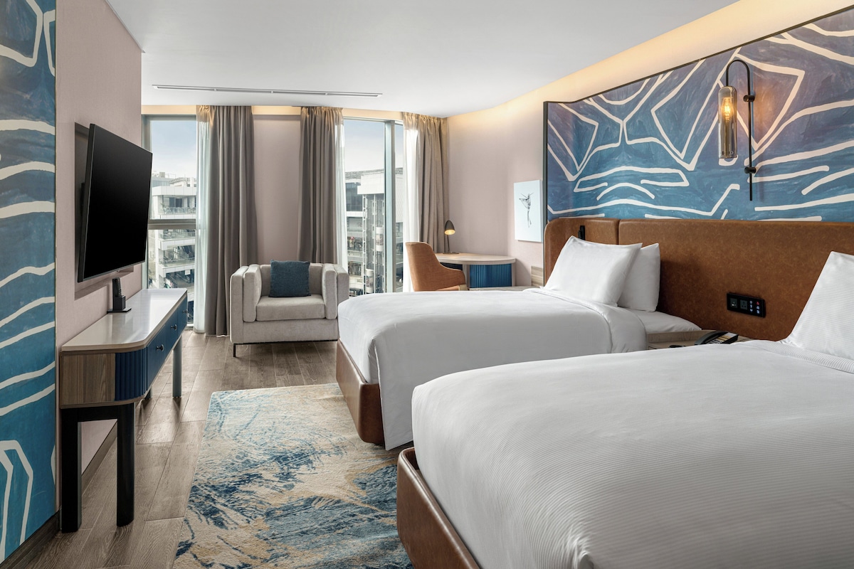Ein Hotelzimmer mit zwei Betten, einem Fernseher, einem Schreibtisch, einem Stuhl, einer Lampe und Blick auf die Stadt.