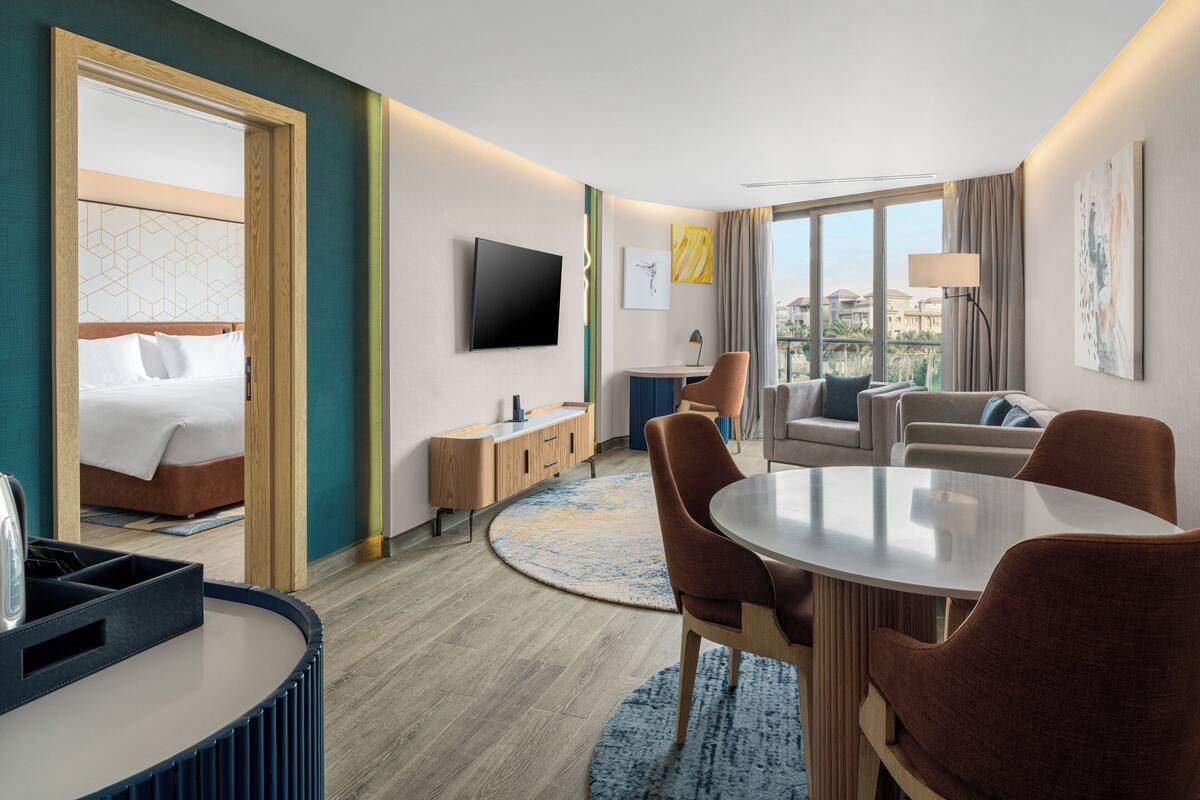 Une chambre d’hôtel comprenant un lit, une télévision, un coin repas avec des chaises, un canapé et une vue sur les bâtiments environnants.
