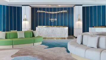 A modern hotel lobby with a reception desk, green and beige sofas, and a blue and white patterned carpet.
