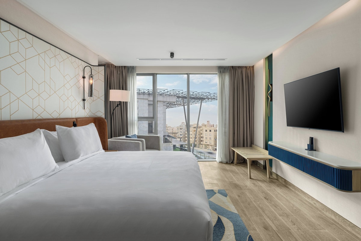 Ein Hotelzimmer mit einem großen Bett, einem Fernseher, einem Schreibtisch und Blick auf die Stadt.