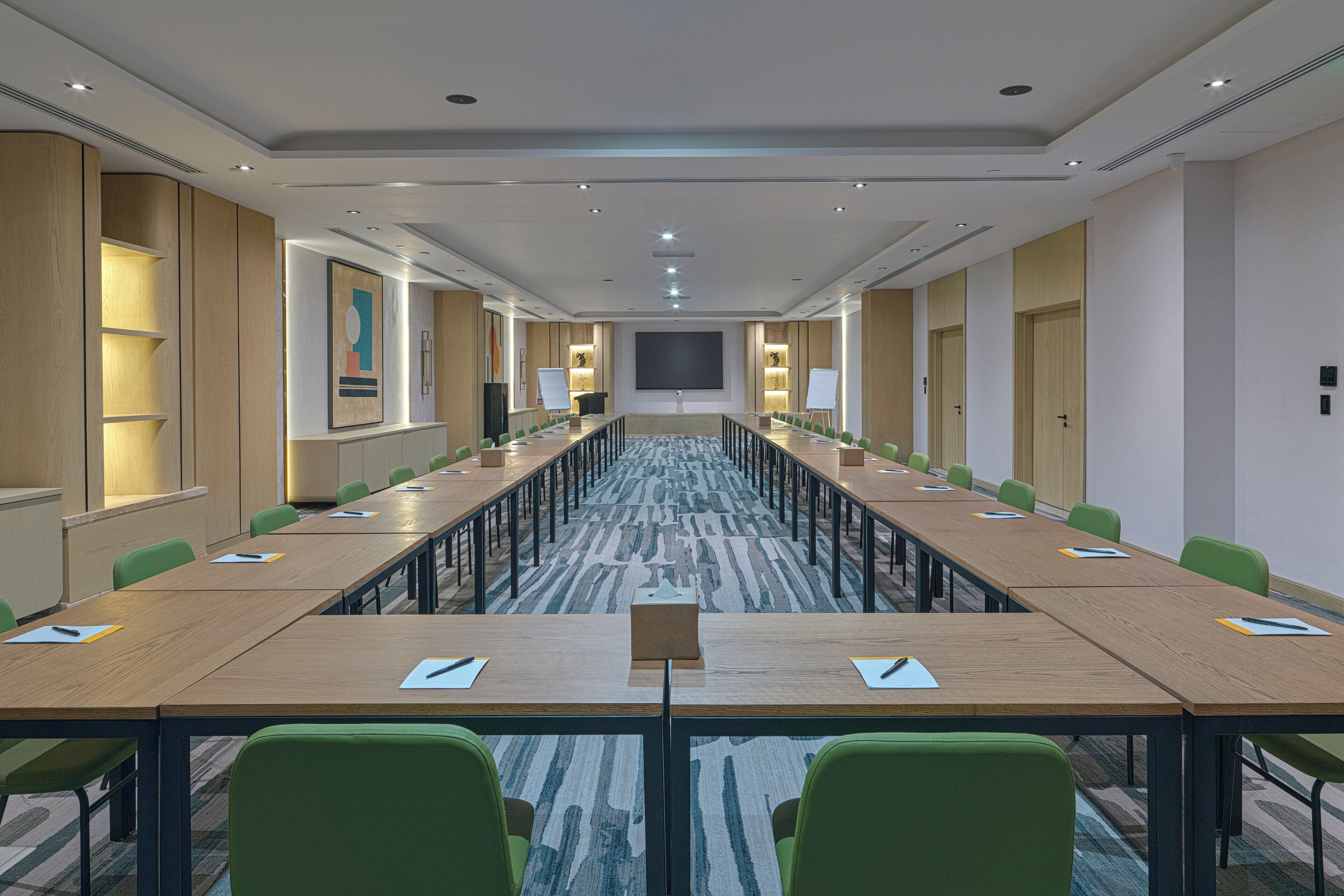 A conference room with long tables, green chairs, a large screen, and a carpeted floor.