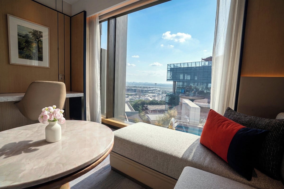 Ein modernes Hotelzimmer mit rundem Tisch, einer Couch mit rotem Kissen, einer Vase mit Blumen, einem gerahmten Bild an der Wand und einem großen Fenster mit Blick auf die Stadt.