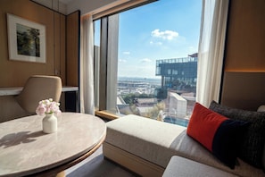 A modern hotel room with a round table, a sofa with a red pillow, a vase with flowers, a framed picture on the wall, and a large window with a city view. - Crowne Plaza Dhaka Airport by IHG (Dhaka)