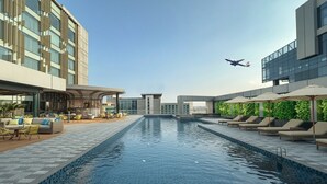 A modern hotel pool area with lounge chairs and umbrellas, a clear sky, and an airplane in the distance. - Crowne Plaza Dhaka Airport by IHG (Dhaka)