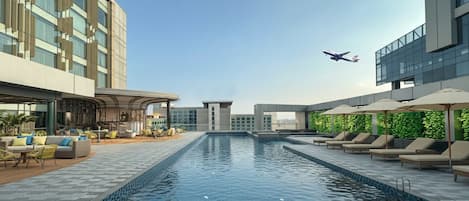 A modern hotel pool area with lounge chairs and umbrellas, a clear sky, and an airplane in the distance.