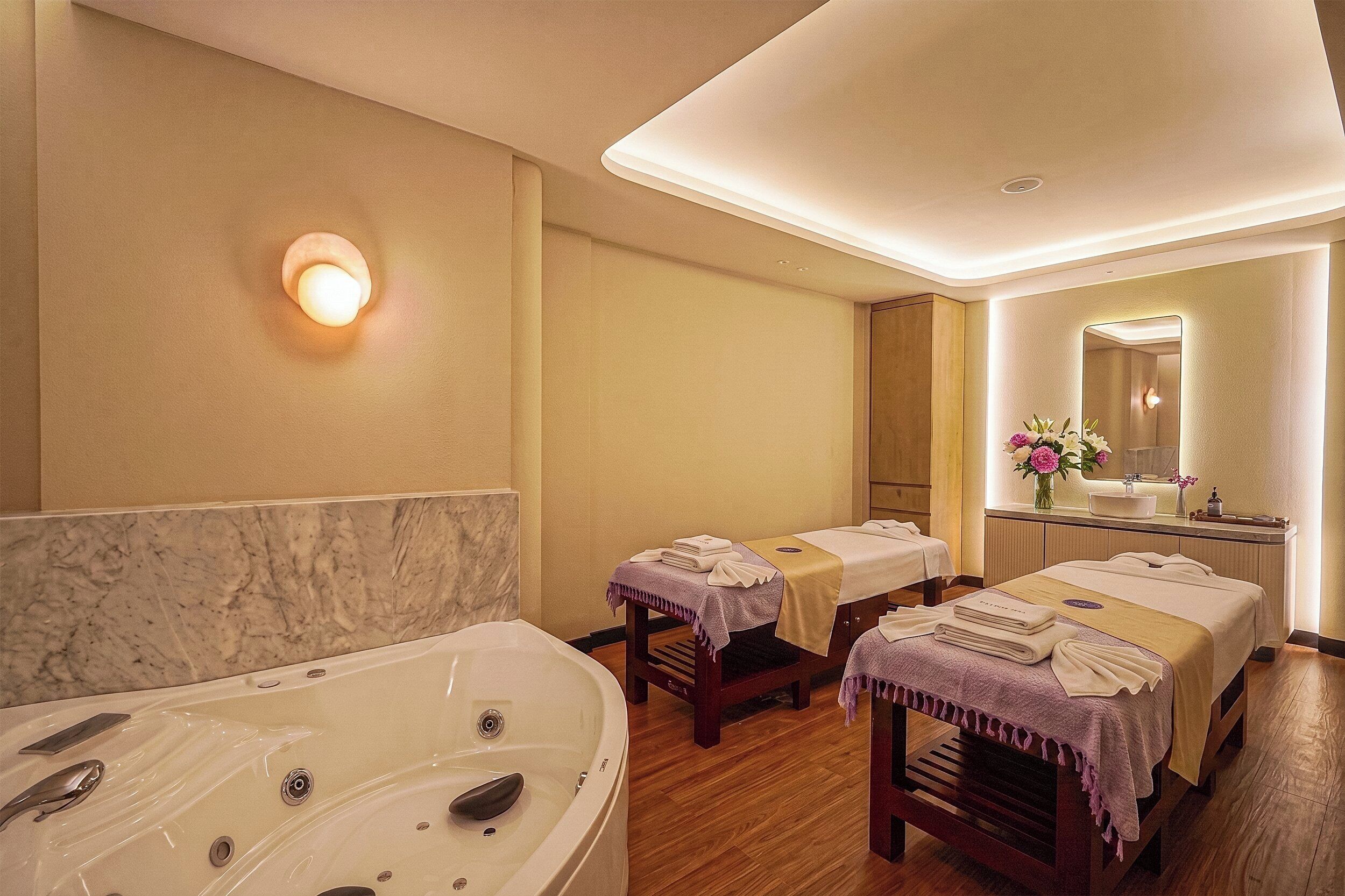 A hotel room with a large bathtub, two beds with towels, a vanity with a mirror, and a wall-mounted light.