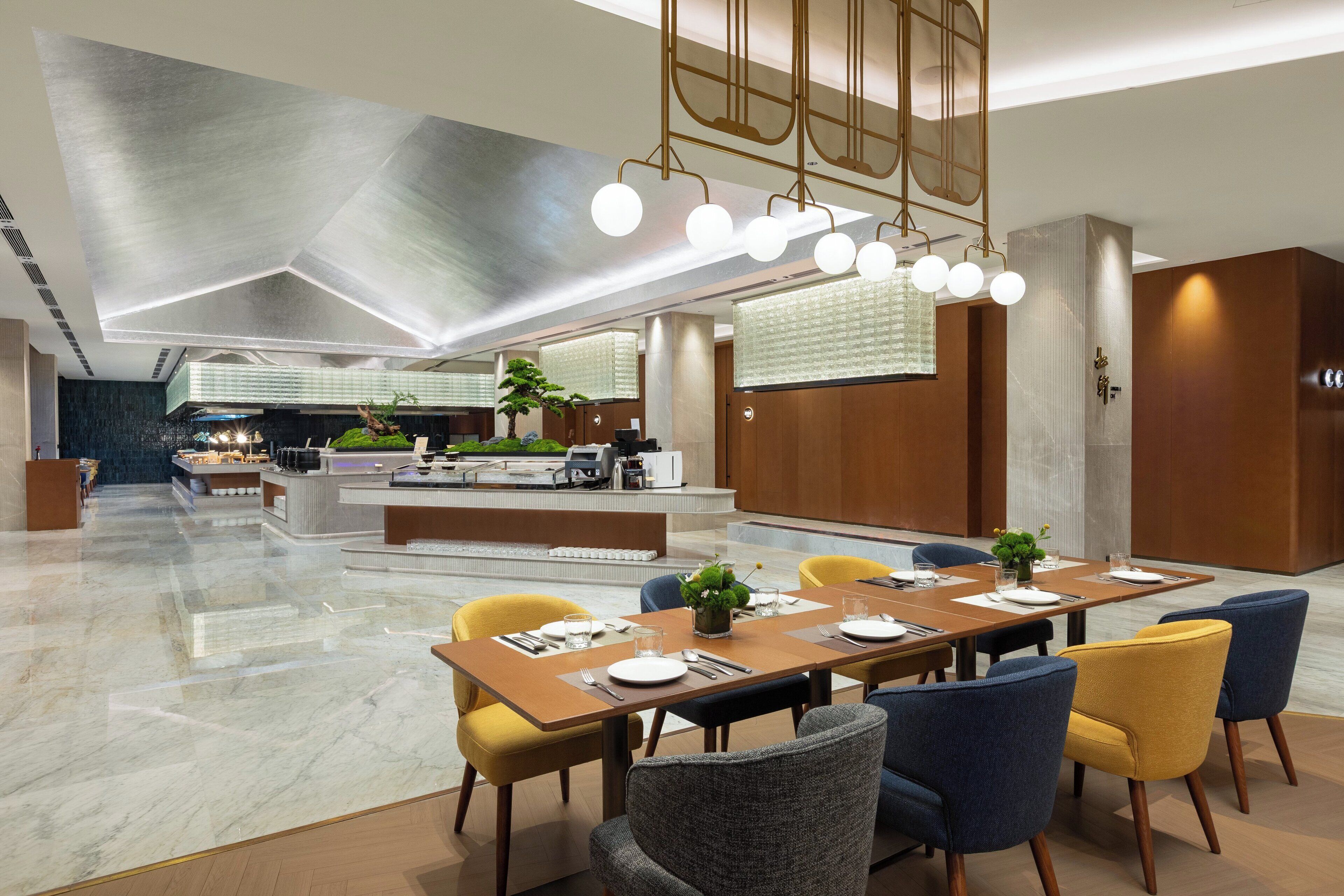 A modern hotel lobby with a long dining table, comfortable seating, and a central reception area.