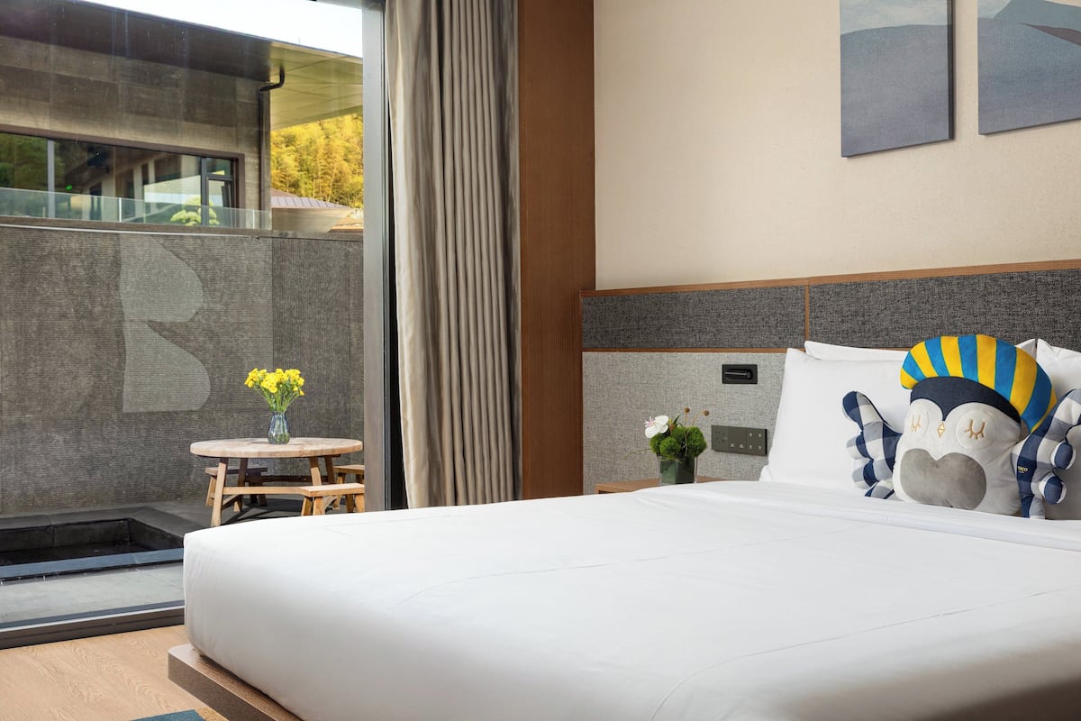 A modern hotel room with a large bed, a small table with flowers, a view of the outdoors, and a unique wall decoration.