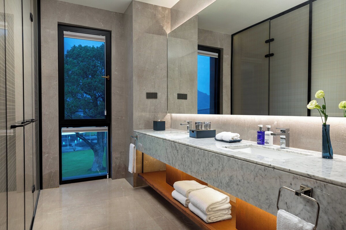A modern bathroom with a large mirror, marble countertop, and a view of a tree outside.