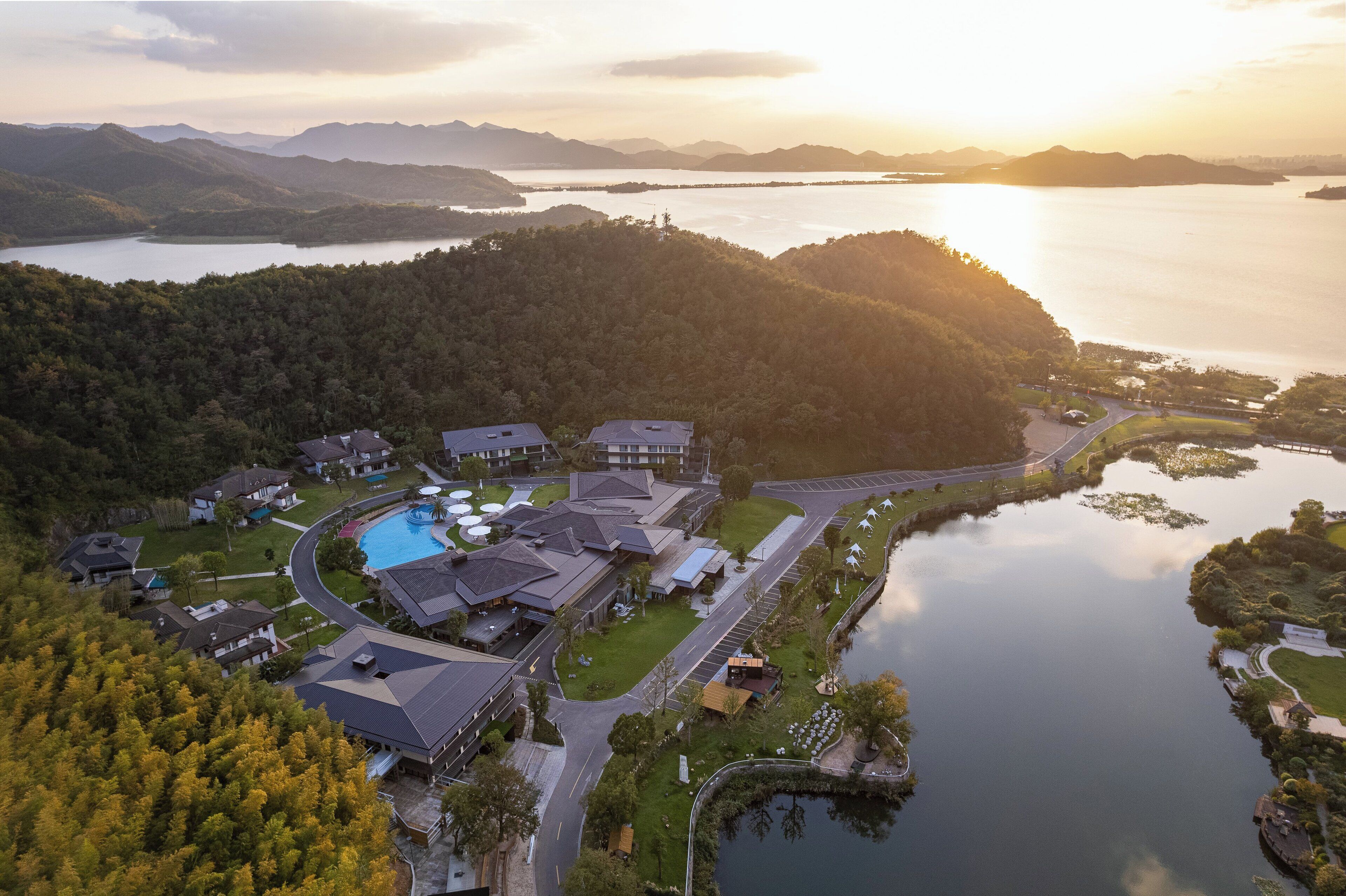 Una vista aerea di un resort con diversi edifici, una piscina e un lago circondato da colline ricoperte di boschi.