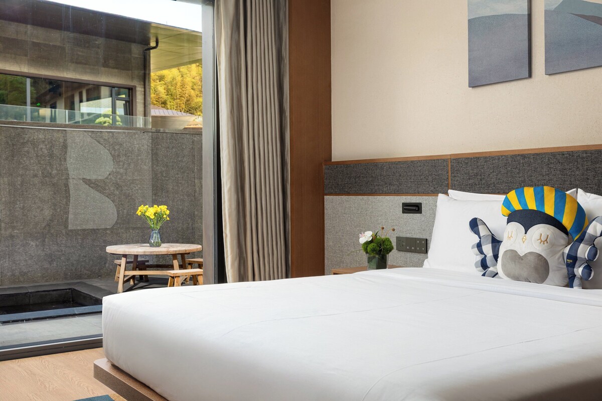 A modern hotel room with a large bed, a small table with flowers, a view of the outdoors, and a unique wall decoration.