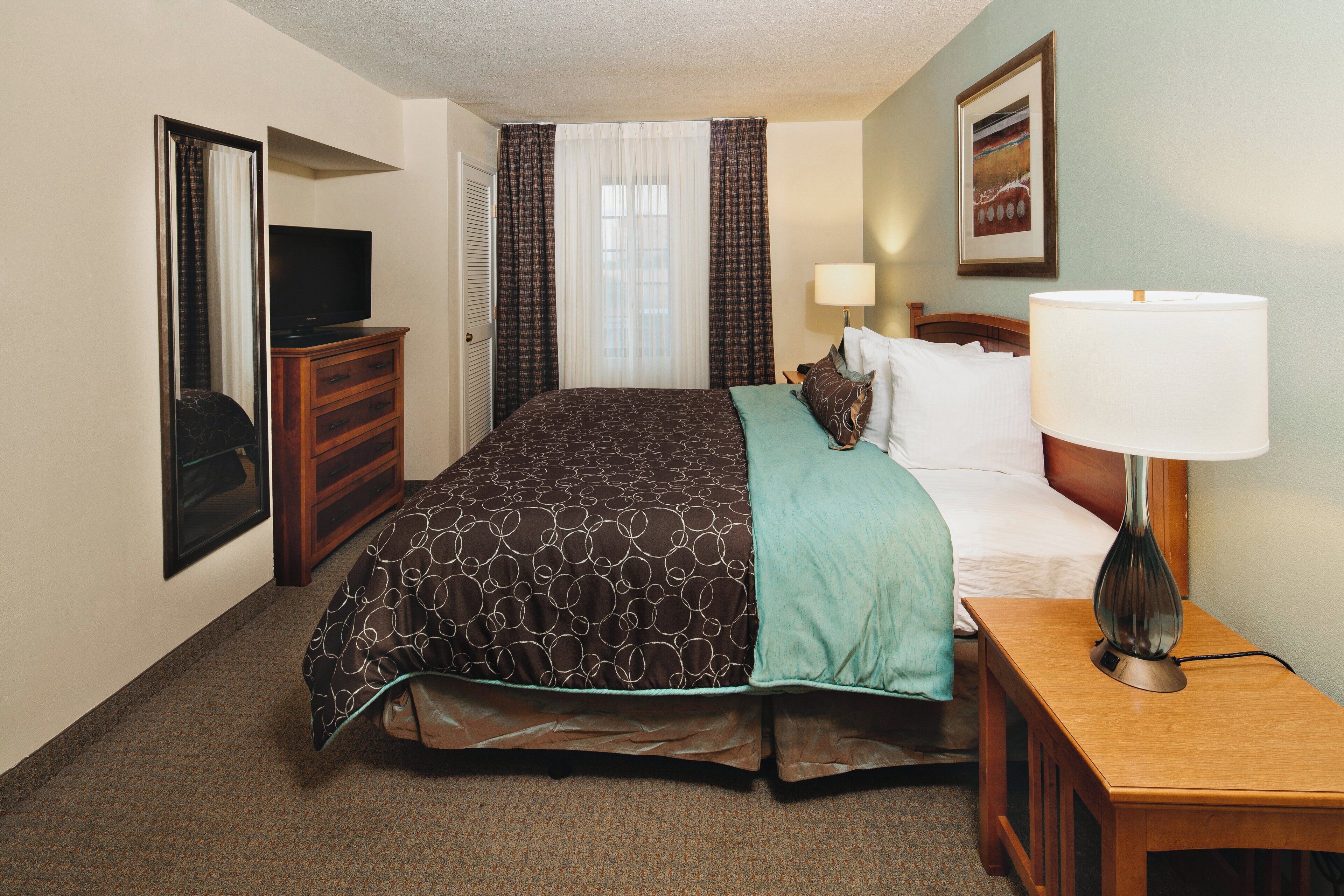A hotel room with a bed, a TV, a dresser, a lamp, and a painting on the wall.
