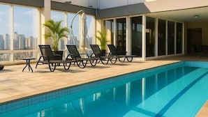 Piscina interior moderna con agua azul cristalina, rodeada de sillones de descanso y vistas a la ciudad.