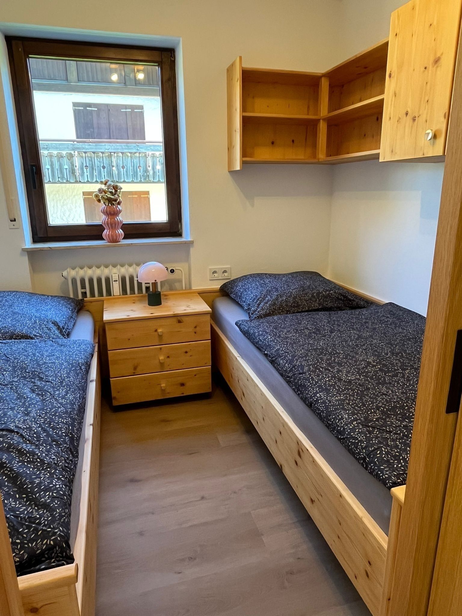 Une petite pièce chaleureuse, avec deux lits en bois, une commode en bois et une fenêtre donnant sur un balcon.
