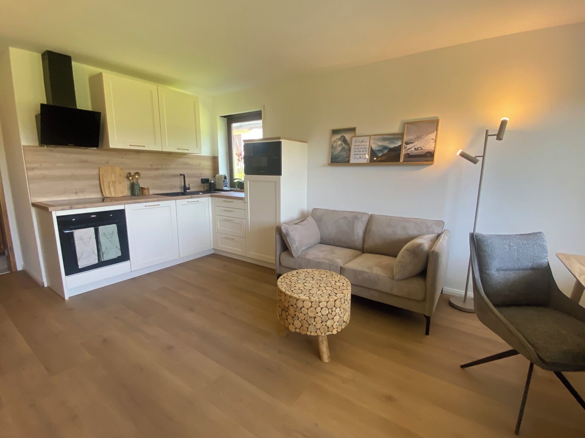 Un espacio habitable pequeño con kitchenette, un sofá, una mesa de centro redonda de madera y una lámpara de pie.