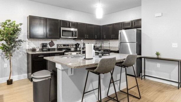 A modern kitchen with dark cabinets, stainless appliances, a granite countertop, and a small island with bar stools. - Grand Prairie 2BR | IKEA, Epic Waters, Bass Pro (Grand Prairie)