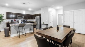 A modern kitchen with dark cabinets, a stainless steel refrigerator, and a wooden dining table with chairs. - Grand Prairie 2BR | IKEA, Epic Waters, Bass Pro (Grand Prairie)