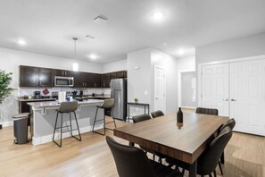 A modern kitchen with dark cabinets, a stainless steel refrigerator, and a wooden dining table with chairs. - Grand Prairie 2BR | IKEA, Epic Waters, Bass Pro (Grand Prairie)