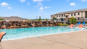 A swimming pool with a waterfall feature, lounge chairs, and a building in the background. - Grand Prairie 2BR | IKEA, Epic Waters, Bass Pro (Grand Prairie)
