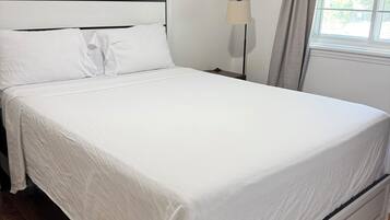 Un llit ben fet en una habitació amb sòl de fusta, una taula de nit amb una làmpada i una finestra amb cortines.