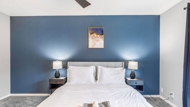 Una camera da letto con un letto, due comodini con lampade, una ventola a soffitto, un quadro appeso al muro e un pavimento con moquette.