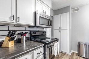 Una cocina con armarios blancos, una estufa negra, un microondas y un cubo de basura de acero inoxidable.