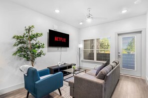A modern living room with a flat-screen TV, a ceiling fan, a large window with blinds, a sofa, a blue armchair, and a potted plant. - Grand Prairie 1BR | IKEA, Epic Waters, Bass Pro (Grand Prairie)