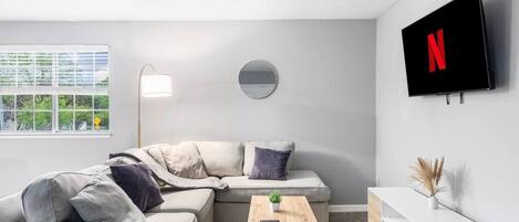 Uma sala de estar moderna com um sofá seccional, um espelho redondo, uma TV montada na parede, uma pequena mesa de madeira e uma janela com vista para o verde.