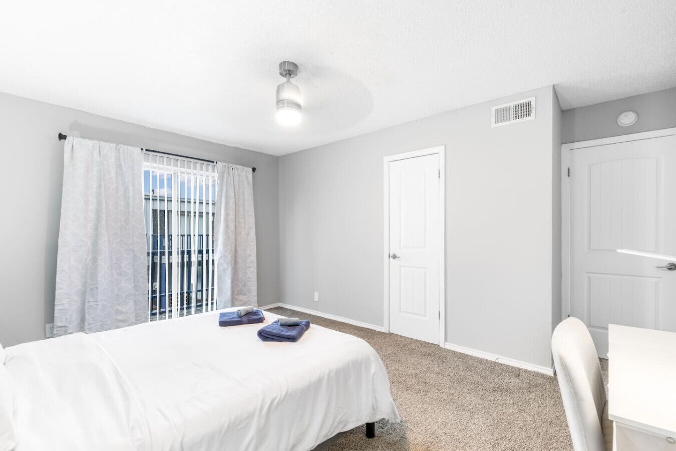 A bedroom with a bed, a desk, a chair, a window with curtains, and a door.