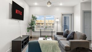 Ein modernes Wohnzimmer mit Flachbildfernseher, Deckenventilator, Fenster mit Jalousien, grauem Sofa und kleinem Tisch mit Pflanze.