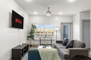 A modern living room with a flat-screen TV, a ceiling fan, a window with blinds, a gray sofa, and a small table with a plant. - Grand Prairie 1BR | IKEA, Epic Waters, Bass Pro (Grand Prairie)