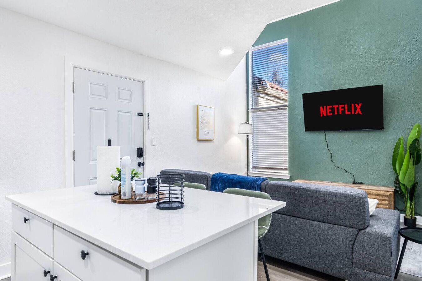 Een moderne woonkamer met een wit bureau, een grijze bank en een tv aan de muur waarop 'NETFLIX' wordt getoond.