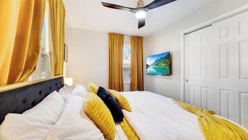 Hotellihuone, jossa on sänky, verhot, kattotuuletin, televisio ja ovi.