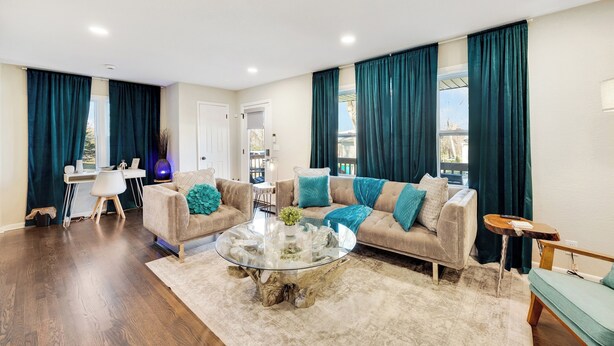 Una sala de estar moderna con una mesa de centro de vidrio, sofás color beige, cojines verde azulado y vistas al exterior a través de amplios ventanales.