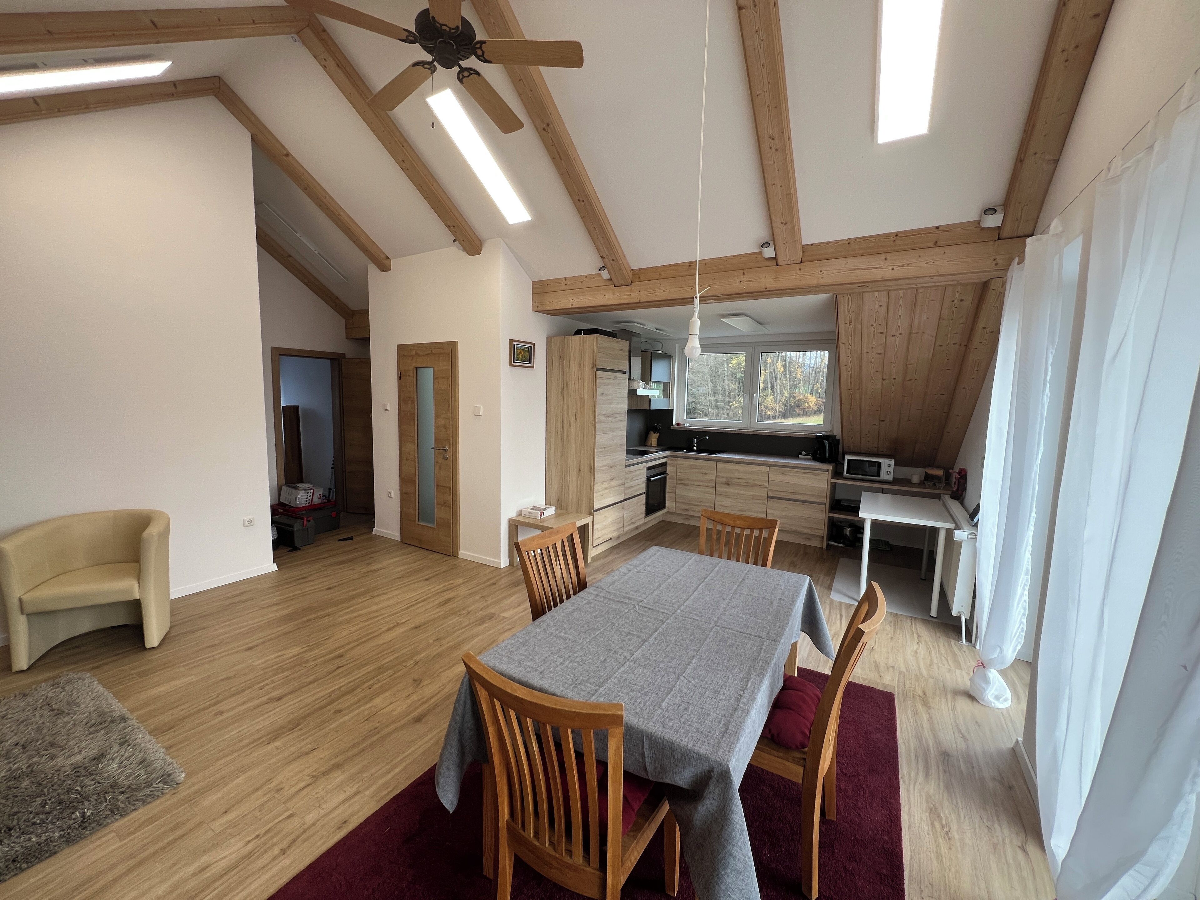 A spacious living area with a dining table, wooden chairs, a ceiling fan, and a kitchen with a window.