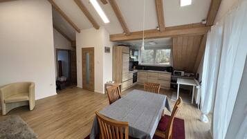 A spacious living area with a dining table, wooden chairs, a ceiling fan, and a kitchen with a window.