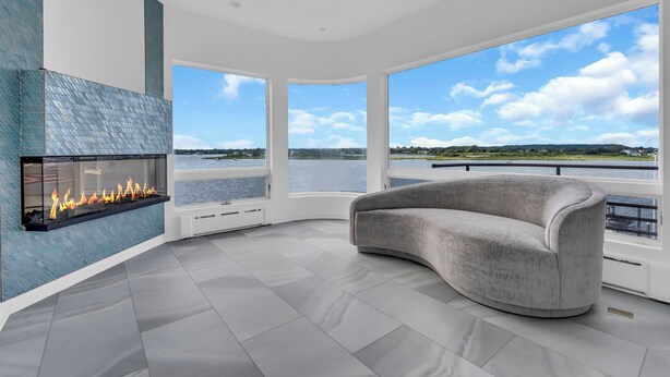 A modern living room with a fireplace, large windows, and a view of a body of water. - Elegant Waterfront Stay + Bay + Sunset Views (Sea Bright)