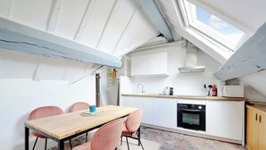 Une cuisine de taille réduite avec des meubles blancs, un four noir et une table en bois accompagnée de chaises roses.