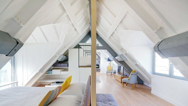 Une chambre mansardée avec de hauts plafonds, comprenant un lit, une banquette et un espace bureau.