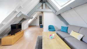 Un appartement moderne de style « loft », avec un plafond en pente, un meuble de rangement en bois pour l’équipement audiovisuel, un canapé gris agrémenté de coussins et une petite table avec des bougies.