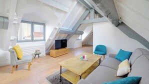 Un salon de style industriel, avec un canapé, un fauteuil et une table basse en bois. Un téléviseur est posé sur un meuble en bois, et une fenêtre laisse entrer la lumière naturelle.