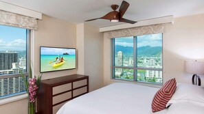Hotelkamer met een bed, een televisie waarop een scene van kajakken te zien is, een plafondventilator en een stadszicht door grote ramen.