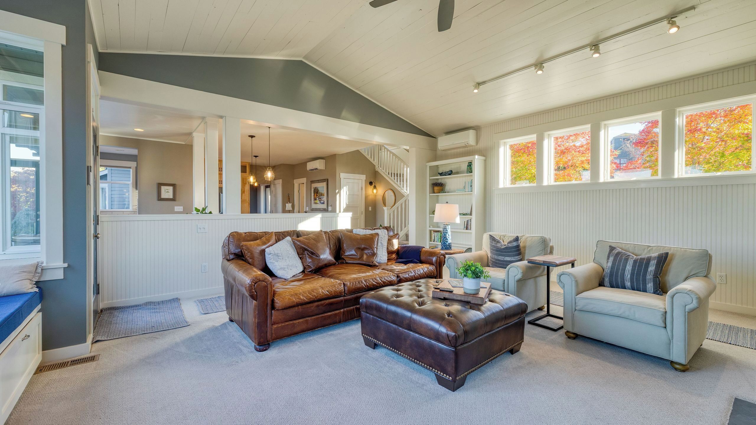 A spacious living room with a brown leather sofa, a light grey armchair, and a matching ottoman. A staircase leads to an upper level, and the room features large windows with autumn foliage views.