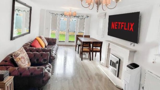 Ein Wohnzimmer mit einer Couch, einem Esstisch, Stühlen, einem Kamin und einem an der Wand befestigten Fernseher, der "NETFLIX" zeigt.