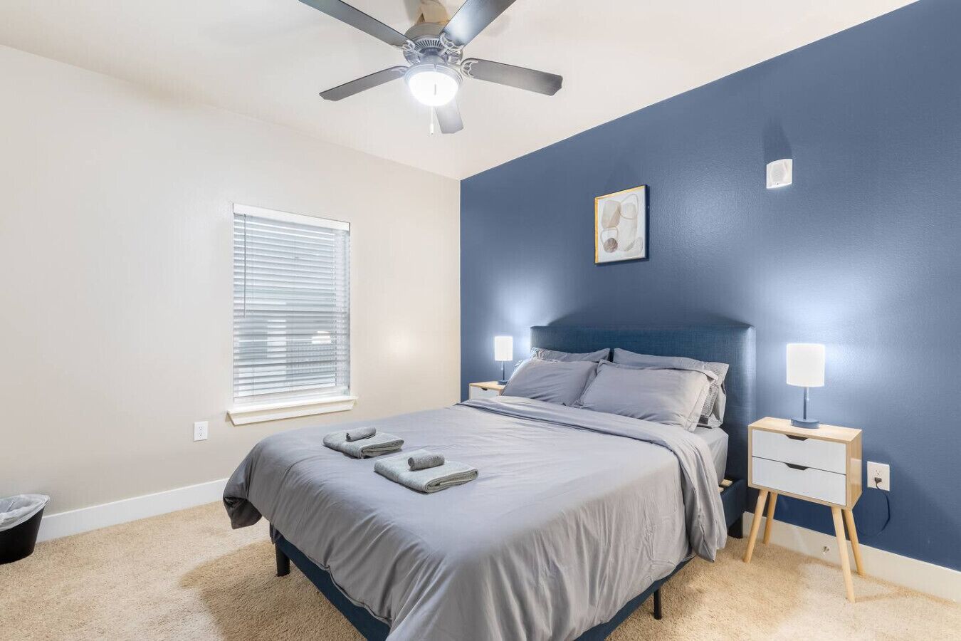 Une chambre à coucher comprenant un lit, des tables de chevet, un ventilateur de plafond, une fenêtre avec des stores et une œuvre d’art accrochée au mur.