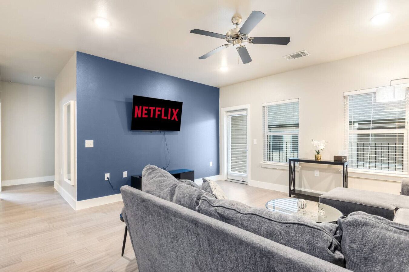 A modern living room with a flat-screen TV mounted on a blue wall, grey sofas, a ceiling fan, and a window with blinds.