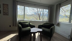 Ett rum med två gröna fåtöljer, ett litet vitt bord och stora fönster med utsikt över träd och berg.